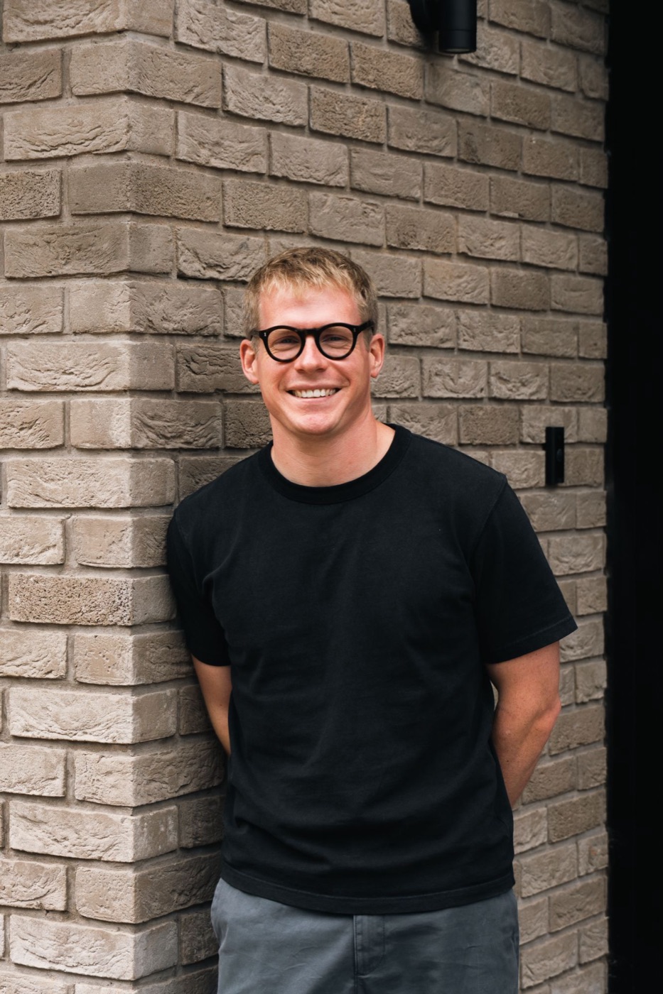 Adam Horton, smiling, leaning against a brick wall in a black T-shirt and round glasses